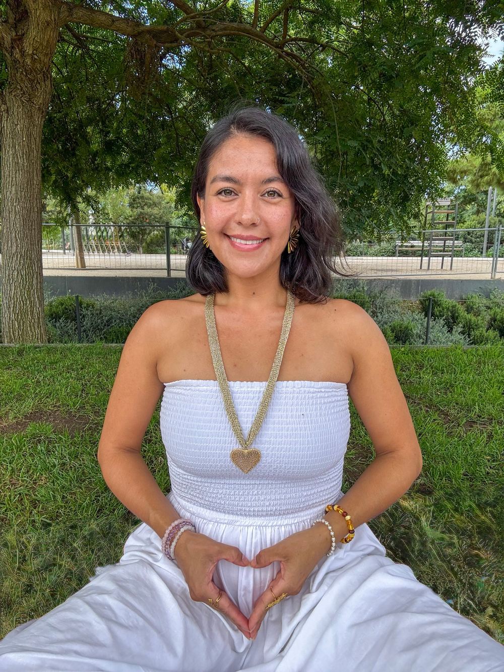 Zayda sonriendo bajo un árbol, vestida de blanco, con las manos formando un corazón